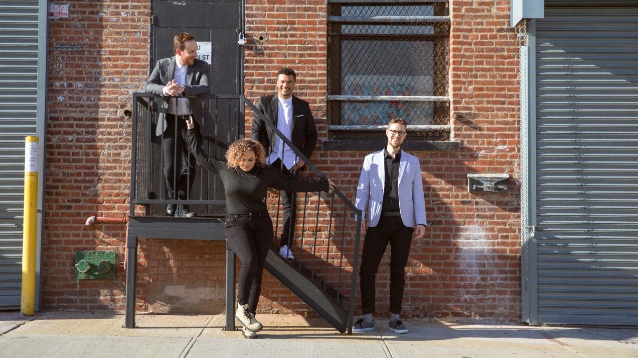  The members of PUBLIQuartet stand on a city sidewalk in front of a brick building with a set of black iron stairs. The female member, a lightskinned Black woman with caramel colored hair stands in front of the stairs and holds onto the railing, dressed in a black turtleneck and black pants. One white man stands on the landing, leaning against the railing in a suit. One man stands on the steps, leaning back up against the brick wall. A third man stands at the bottom of the steps in a beige blazer and dark pants.