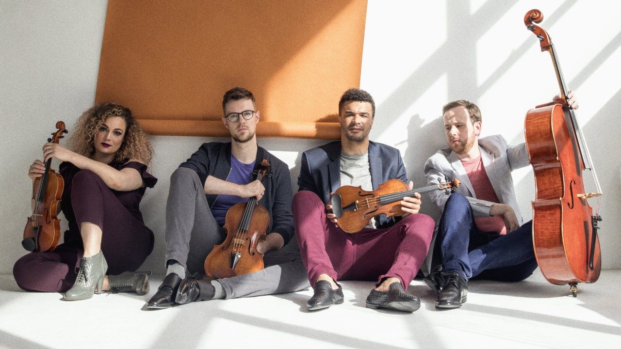  The members of PUBLIQuartet sit on the floor of a photo studio holding their instruments. The room is painted white and a terracotta-colored roll of butcher paper hangs down the wall behind them. The group is, from left, a light skinned Black woman with caramel colored natural hair, a white man with glasses, a light skinned Black man, and a whtie man with a reddish brown closely trimmed beard.