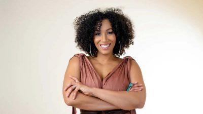  Country musician Rissi Palmer, a light-skinned Black woman with curly brown hair, wears a a brown dress, nude lipstick, large thin gold hoops, and a large turquoise ring and smiles towards the camera with her arms crossed.