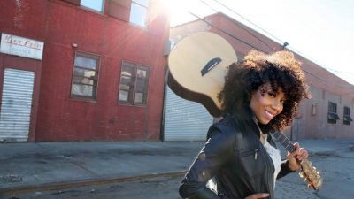  Country musician Rissi Palmer, a light-skinned Black woman with curly brown hair, wears a black leather jacket and a white shirt, and stands in the street in front of run down red brick commercial buildings. She holds an acoustic guitar by the neck and props the body up on her left shoulder.