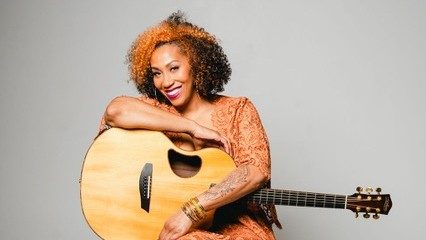  Country musician Rissi Palmer, a light-skinned Black woman with curly brown and blonde hair, holds an acoustic guitar in her lap and wears an orange dress, magenta lipstick, and large thin gold hoops, smiling towards the camera.