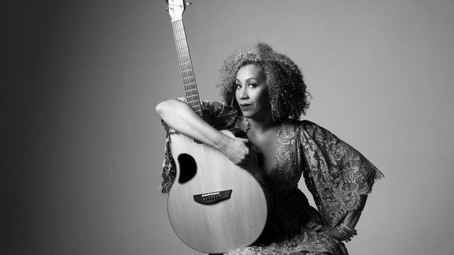  Country musician Rissi Palmer, a light-skinned Black woman with curly brown and blonde hair, holds an acoustic guitar in her lap and wears an orange dress, magenta lipstick, and large thin gold hoops, smiling towards the camera in this black and white image.