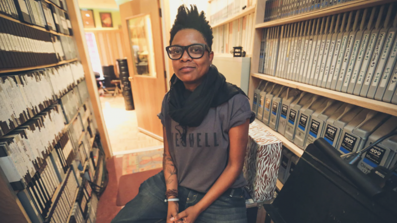  Shirlette Ammons, a Black woman with natural hair cut into a fade, sits on  stool in a library row wearing jeans, a grey t-shirt with rolled sleeves, black scarf, and black framed glasses. She smiles lightly at the camera.