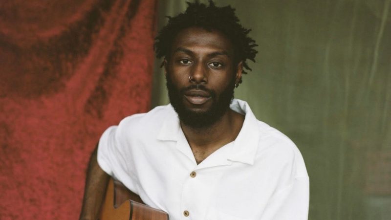  Sonny Miles, a Black man with medium length twists and a full beard, wears a white button down t-shirt and a silver hoop nose ring in one nostril. He sits and holds an acoustic guitar. Behind him are draped two pieces of fabric, one heavier, possibly red velvet, and one thinner, a light green sheer fabric. He looks towards the camera with a neutral expression, lips slightly parted.