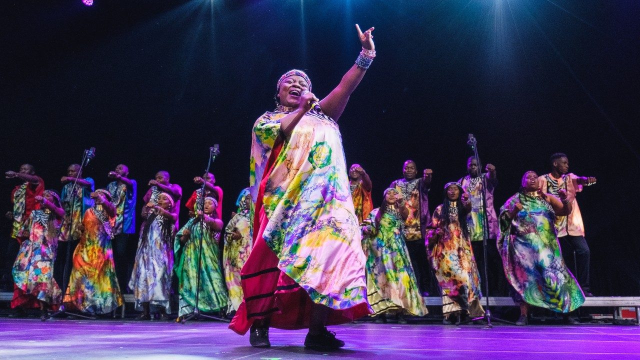  Members of Soweto Gospel Choir perform onstage in colorful traditional costumes. In the foreground, a Black woman sings into the microphone, both eyes closed, her left pointer finger high above her head.