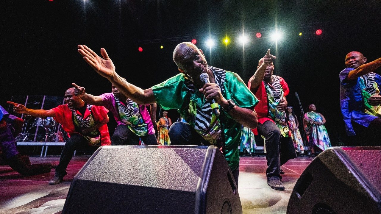  Members of Soweto Gospel Choir perform onstage in colorful traditional costumes. In the foreground, the men line up and point towards the audience. In the very front, a Black man in green sings into the microphone, his right hand extended towards the audience.