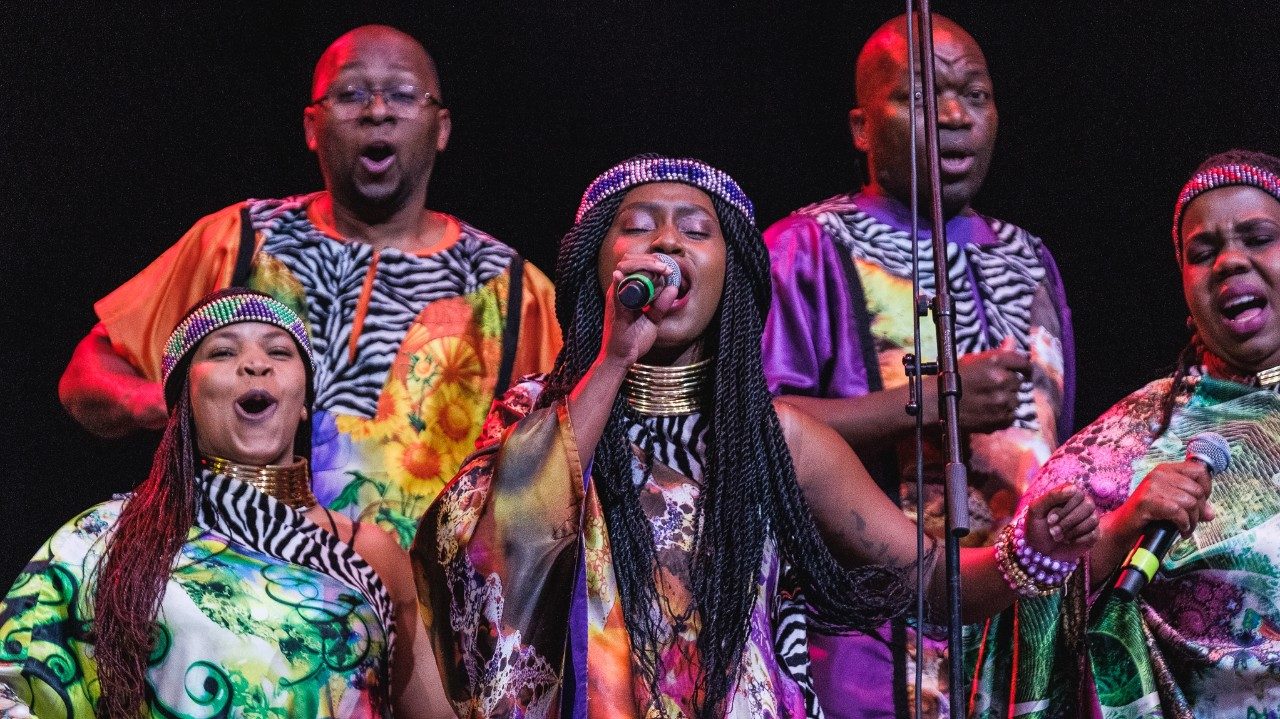  Members of Soweto Gospel Choir perform onstage in colorful traditional costumes. Front and center, a Black woman with long dark brown twists and a purple headband sings into the micrphone with her eyes closed.