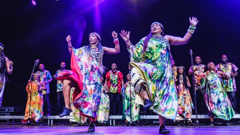  Members of Soweto Gospel Choir perform onstage in colorful traditional costumes. In the foreground, two Black women with long braids and twists dance, arms extended to both sides like a cactus, right leg bent at a 90 degree angle, thighs parallel to the floor.