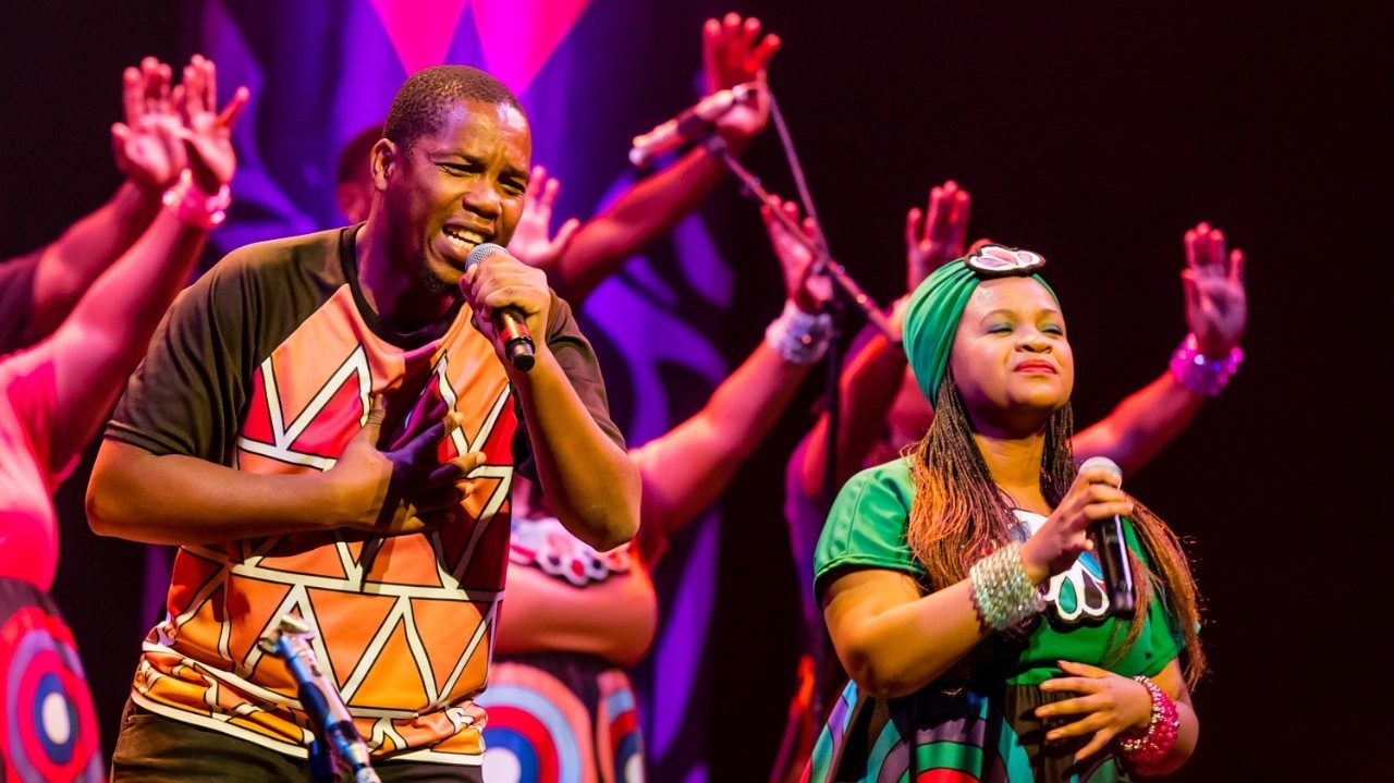  Members of Soweto Gospel Choir sing during a performance. On the left is a Black man singing into a microphone and touching his chest. On the right is a Black woman with a green head wrap, long light brown braids, and traditional costume.