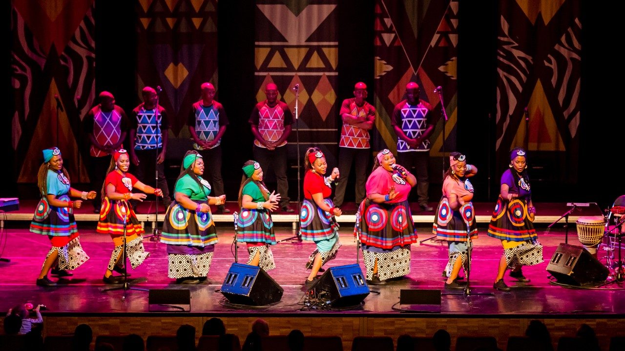  Members of Soweto Gospel Choir perform onstage, the women all in a line facing to their left in the foreground, and the men all in a line behind them. They all wear colorful traditional costumes.