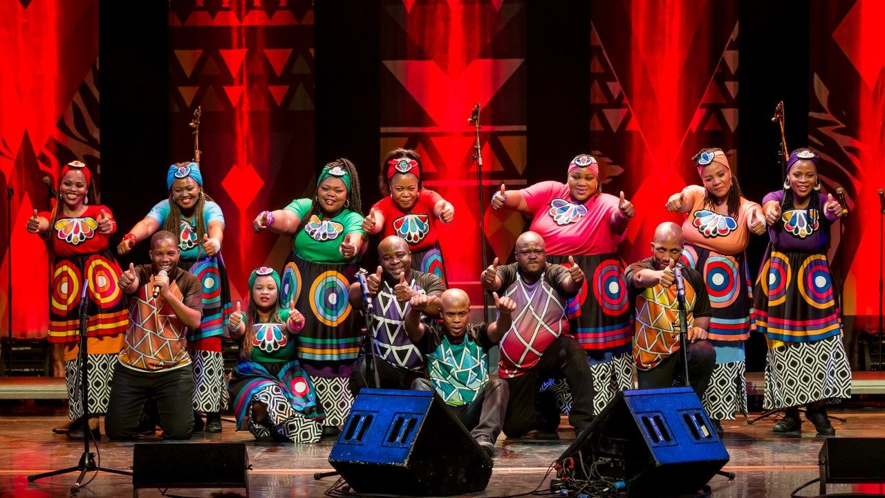  Members of Soweto Gospel Choir pose onstage, everyone giving thumbs up with both hands towards the camera.