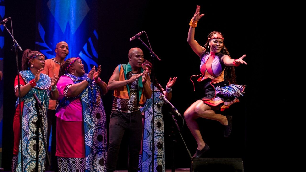  Members of Soweto Gospel Choir perform onstage, all in bright traditional costumes. In the background, members sing and clap along. In the foreground, a Black woman with long light brown braids, jumps into the air, her legs bent beneath her. She reaches her left arm out straight towards the audience, her right arm up above her head.