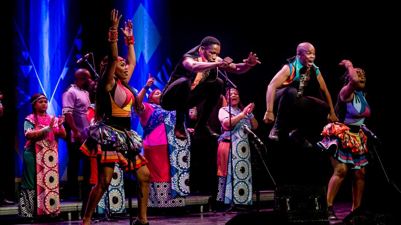  Members of Soweto Gospel Choir perform onstage in colorful traditional costumes. Some sing and clap. Two Black men, in the foreground, tuck jump high into the air.