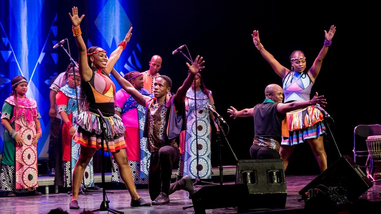  Members of Soweto Gospel Choir perform onstage, some singing in the background. Others in the foreground pose with both arms raised high above their heads in a V shape.