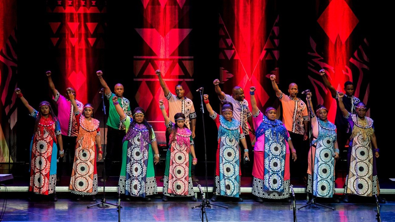  Members of Soweto Gospel Choir perform onstage in colorful traditional costumes and all raise their right fists high into the air above their heads.