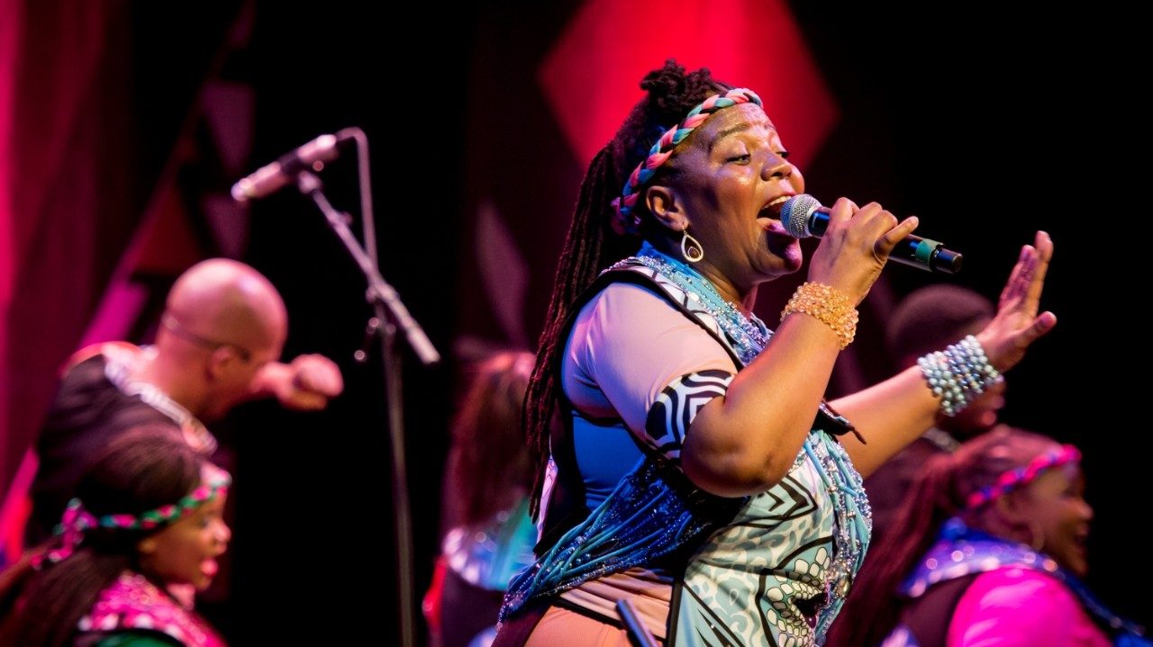  Members of Soweto Gospel Choir perform onstage in colorful traditional costumes. In the foreground, a Black woman with long braids pulled into a ponytail sings into a microphone, her other hand extended out and away from her.