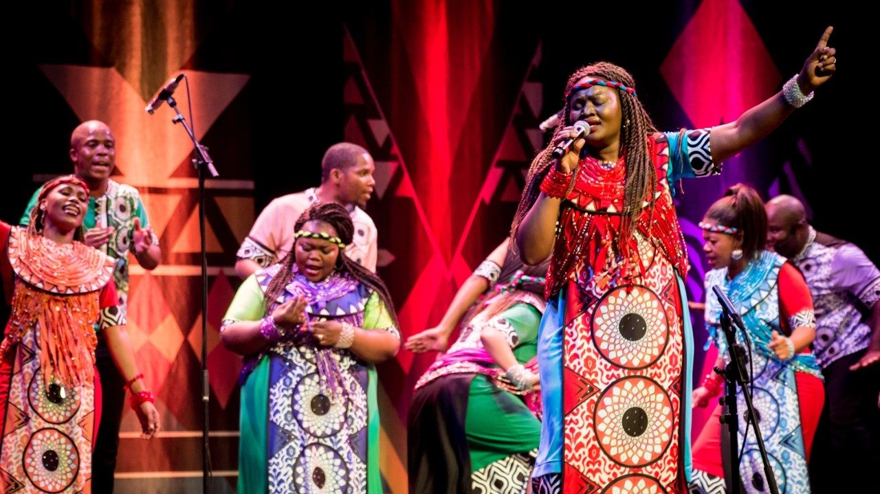  Members of Soweto Gospel Choir perform onstage in colorful traditional costumes. In the foreground, a Black woman with long braids sings into the microphone with her eyes closed, left arm raised above her head with her index finger extended.