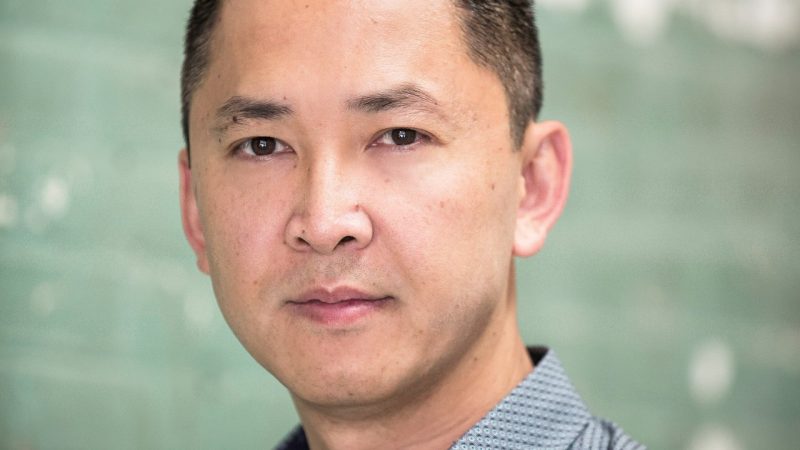  Author Viet Thanh Nguyen, an Asian man with dark brown hair cut long on top and with a fade on the sides, wears a grey patterened button down shirt in front of a sea glass green background.