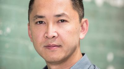  Author Viet Thanh Nguyen, an Asian man with dark brown hair cut long on top and with a fade on the sides, wears a grey patterened button down shirt in front of a sea glass green background.