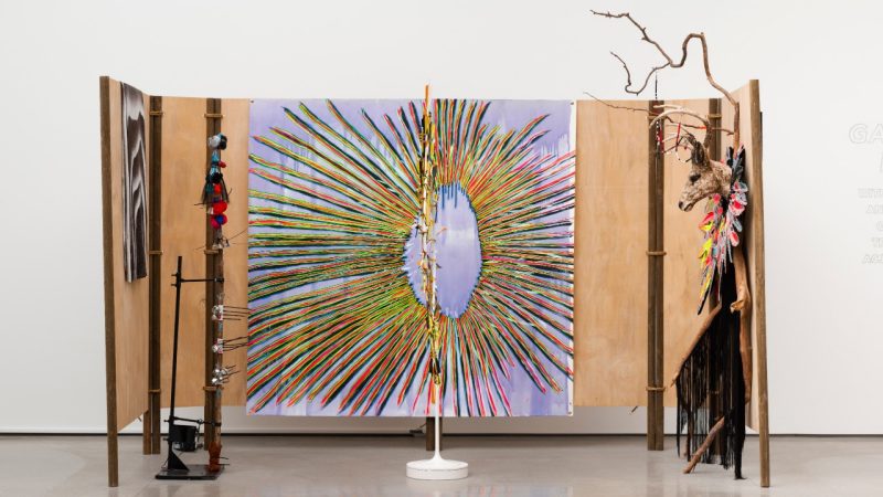  An installation view of Laurie Steelink's "Gathering Power," 2022. Photo by Yubo Dong, ofstudio. A three-paneled wood structure on which hangs two paintings. The painting in the center is a multicolored starburst or dream catcher pattern. Sculptural elements at left and in the center are reminiscent of totem poles. At right, a deer head and a willowy tree branch hang on a panel.