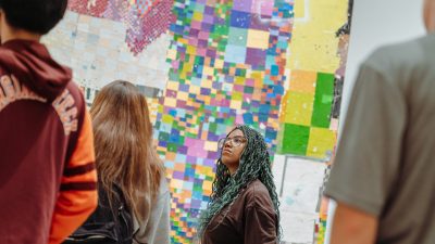 Virginia Tech students visit the Moss Arts Center galleries to view the fall 2023 exhibitions. At center, a young Black woman in a brown shirt, wearing glasses and with blue woven into her long braids, observes william cordova's exhibition.