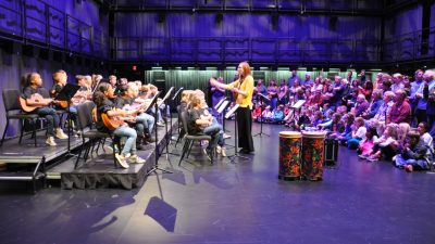 The Belview Ukulele Club performs in the Cube at the Moss Arts Center, led by music teacher Natalie Gibbs (center), a white woman with red hair wearing a yellow sweater and black pants.