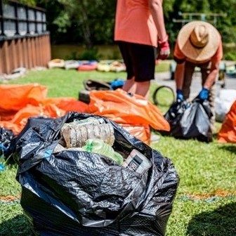 Two people in orange T-shirts pick up litter in the background. In the foreground is a black plastic garbage bag full of collected litter.