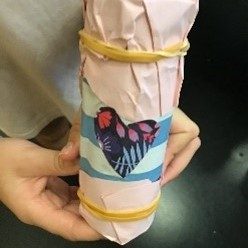 A child's hands hold a homemade shaker, decorated with construction paper and secured with two rubber bands. A heart cut out of floral paper is on the front.