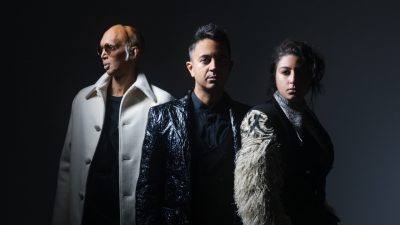  From left, Shahzad Ismaily, a bald brown man with white sideburns, round sunglasses, and a long white peacoat over a dark shirt; Vijay Iyer, a young brown man with medium length dark brown hair in a fade on the sides, wearing a black and silver brocade blazer over a black button down shirt, and Arooj Aftab, a young brown woman with dark brown hair pulled into a messy updo, wearing a black jacket with fluffy white sleeves. They stand in front of a dark grey background.