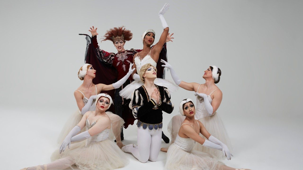  Seven of the dancers of Les Ballets Trockadero de Monte Carlo are dressed as characters from Swan Lake. Five are dressed in all white as swans. One, in the center, is dressed as a prince and kneels, palms pressed together in a prayer pose. The last, in the back, is dressed as an evil sorceress with wild, unkempt red hair. All seven are men dressed in beautiful, detailed drag.