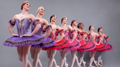  Eight of the dancers from Les Ballets Trockadero de Monte Carlo, all men dressed in beautiful, detailed drag. They wear tutus in shades of pink, purple, and red, and stand in a line, all in the same position with hands on their hips and looking towards the top right edge of the frame.