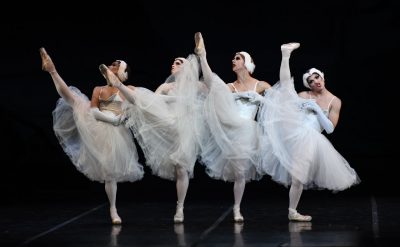 Members of all-male comic ballet group Las Ballets Trockadero de Montecarlo wear white tutus and dance on stage, one dancer pulling a funny face, as they all kick their right legs high into the air.