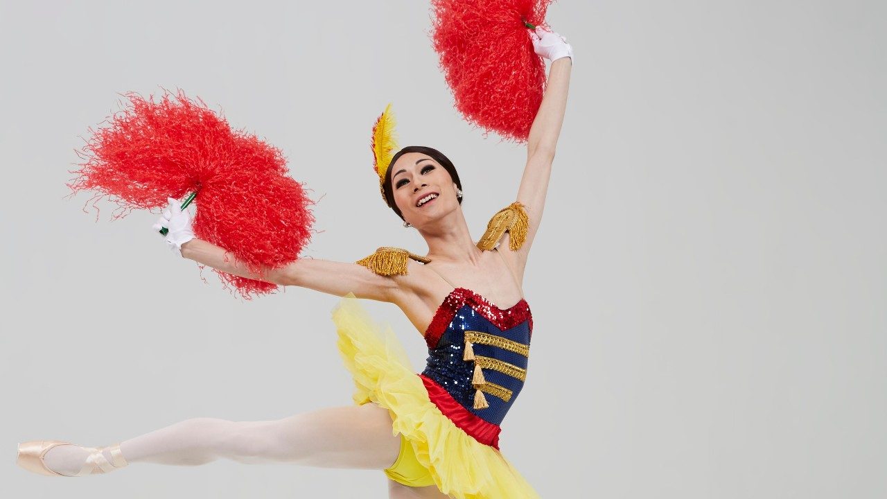  A dancer from Les Ballets Trockadero de Monte Carlo, a man in drag holding two red feather fans and a stars and stripes inspired uniform with a yellow tutu. He stands en pointe, his right leg extended to the side and arms raised.