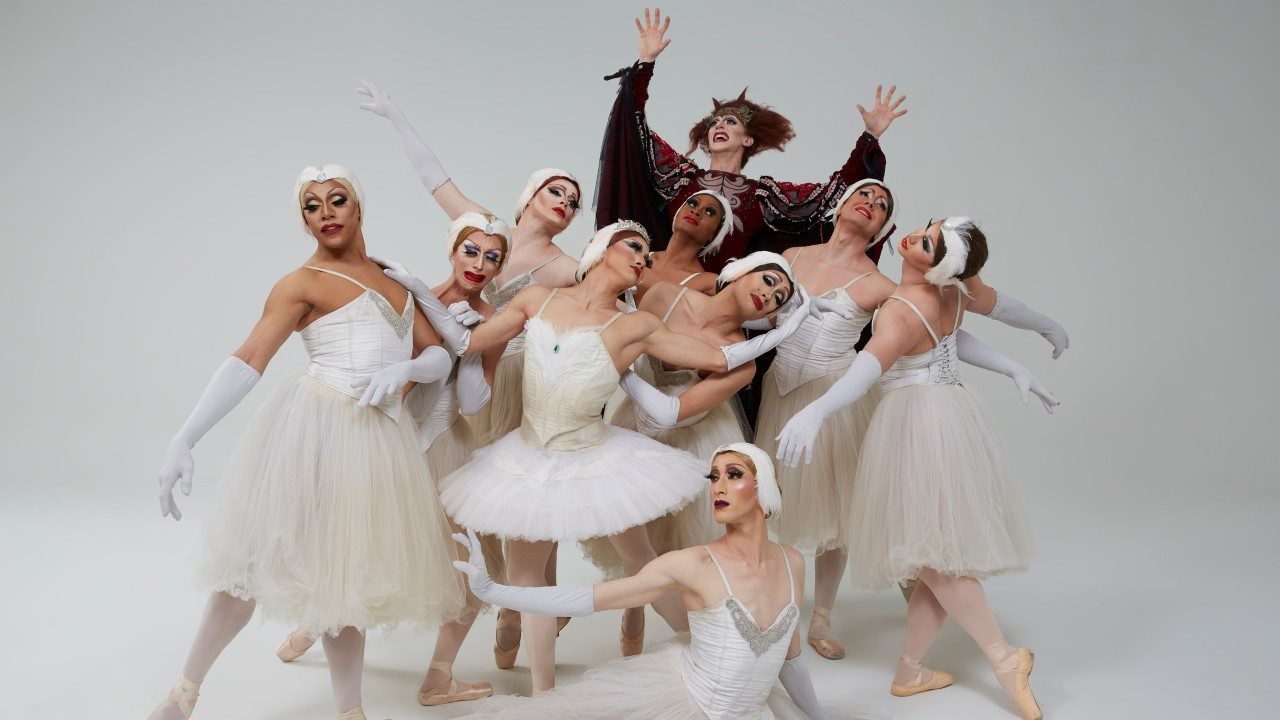  The dancers of Les Ballets Trockadero de Monte Carlo wear extravagant costumes. Nine of the ten dancers wear white tutus and feathered head pieces, while the in the back, the tenth dancer is dressed as the evil sorceress. All 10 are men dressed in beautiful, detailed drag.
