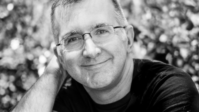  Music critic Alex Ross, a white man with short salt and pepper hair and frameless rectangular glasses, wears a black T-shirt and smiles towards the camera outside under dappled sunlight in this black and white image.