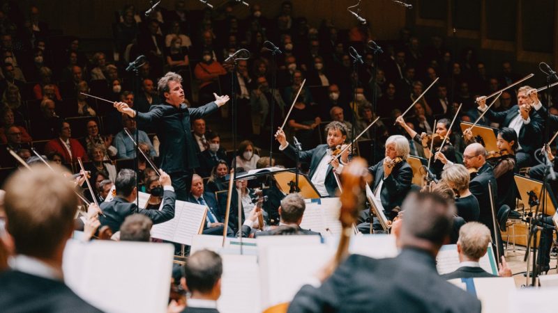 Conductor Jakub Hrůša, a middle aged white man with sandy brown hair, extends his arms out wide as he conducts the Bamberg Symphony during a live performance. All the string players watch him and their bows are raised in the air away from their instruments.