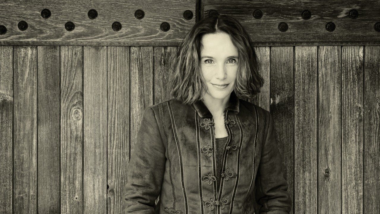  Pianist Hélène Grimaud, a middle aged white woman with chin-length wavy hair wears a jacket and smiles towards the camera, a wooden fence behind her, in this black and white photo.