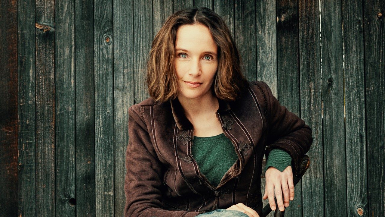  Pianist Hélène Grimaud, a middle aged white woman with chin-length wavy brown hair and blue eyes wears a brown jacket and forest green shirt and smiles towards the camera, a wooden fence behind her.