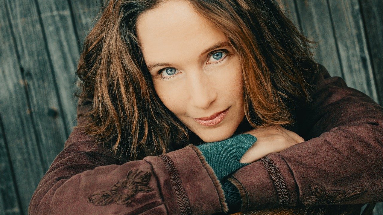  Pianist Hélène Grimaud, a middle aged white woman with chin-length wavy brown hair and blue eyes wears a brown jacket and forest green shirt. She rests her chin on her crossed arms and smiles towards the camera, a wooden fence behind her.