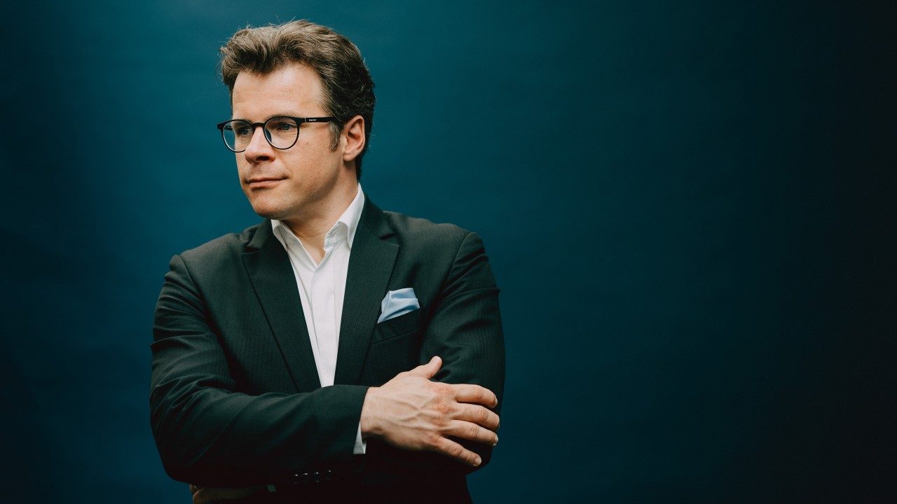  Conductor Jakub Hrůša, a middle aged white man with sandy brown hair and black, round framed glasses, wears a white button down shirt, black jacket, and baby blue pocket square in front of a deep teal background. He crosses his arms across his body and smiles lightly as he looks towards the left edge of the frame.