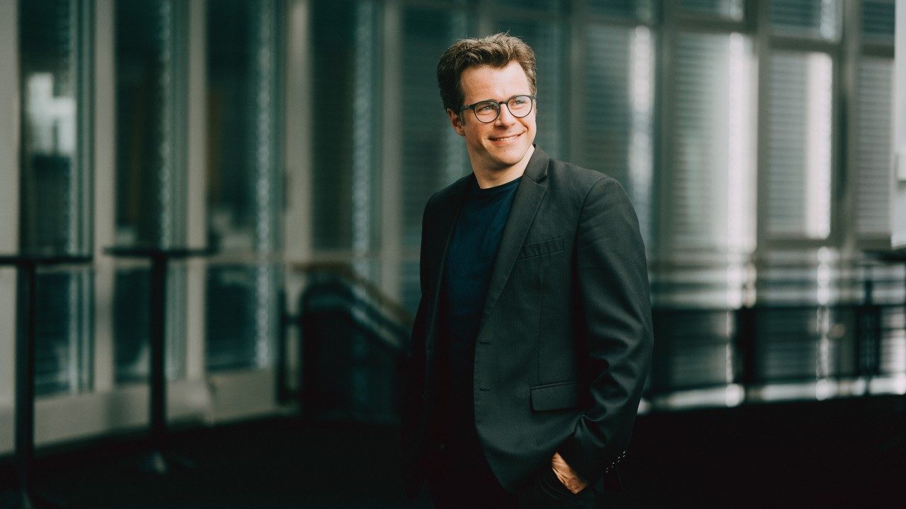  Conductor Jakub Hrůša, a middle aged white man with sandy brown hair and black, round framed glasses, wears a black T-shirt under a black suit with his hands in his pockets. He grins widely as he looks towards the right edge of the frame. Behind him is a series of industrial windows with mini blinds and a black metal railing.