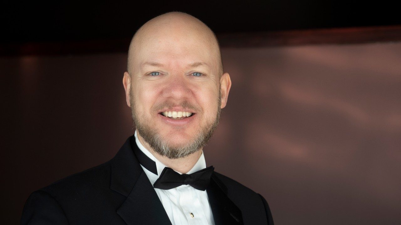  A headshot of Dwight Bigler, the conductor of the Blacksburg Master Chorale. Bigler is a middle aged white man with a bald head and closely trimmed greying blonde beard. He wears a tuxedo.
