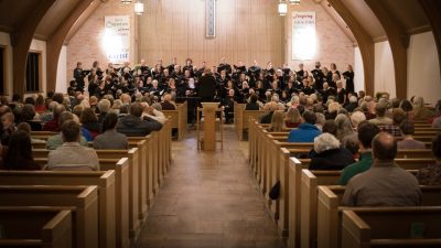  Blacksburg Master Chorale performs in a church, the photo taken from the back between the pews as people watch.