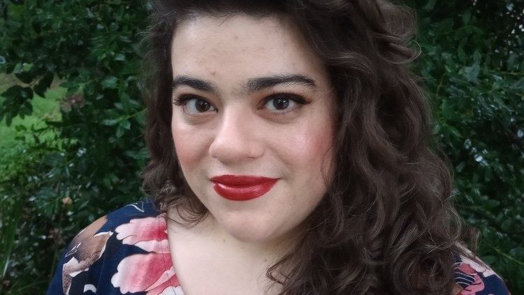 Mezzo-soprano Kayla Brotherton, a white woman with dark, long, wavy brown hair and brown eyes wearing red lipstick and a blue and pink floral shirt. She stands in front of  greenery and looks towards the camera.