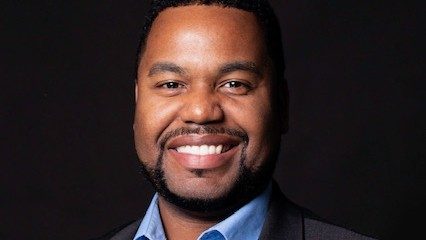 Bass-baritone Markel Williams, a Black man with a dark brown goatee and wearing a dark blazer over a light blue button down shirt, smiles towards the camera in front of a dark background.