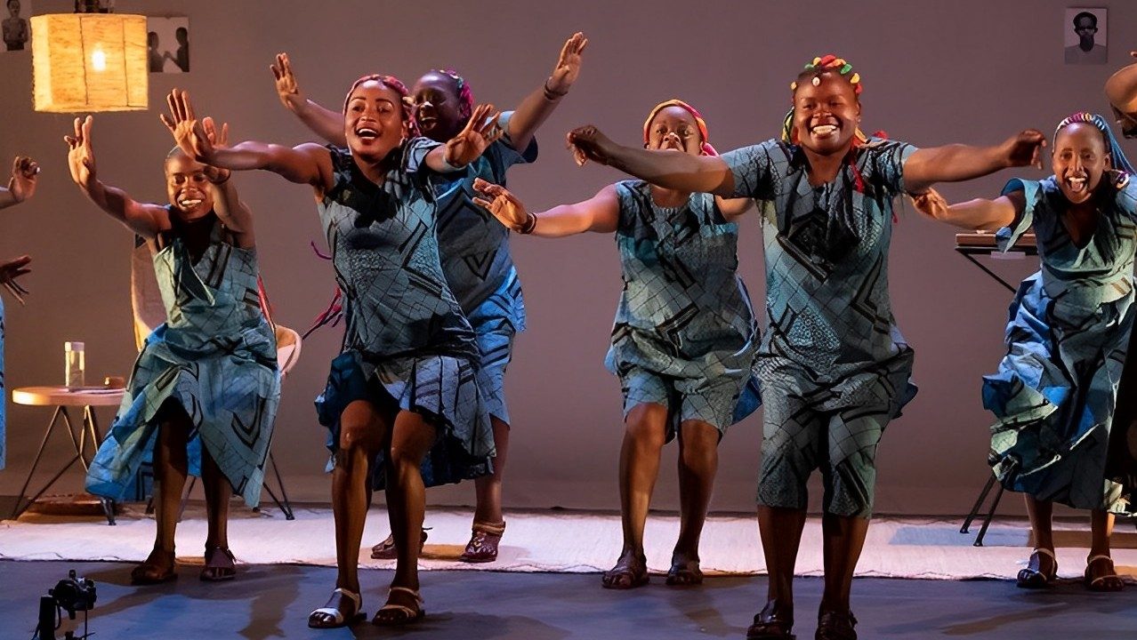  Women jump and dance on stage during a performance of "The Book of Life." The women are Black with brightly colored long braids, and they all wear blue and brown patterned dresses of varying styles but all made from the same fabric.