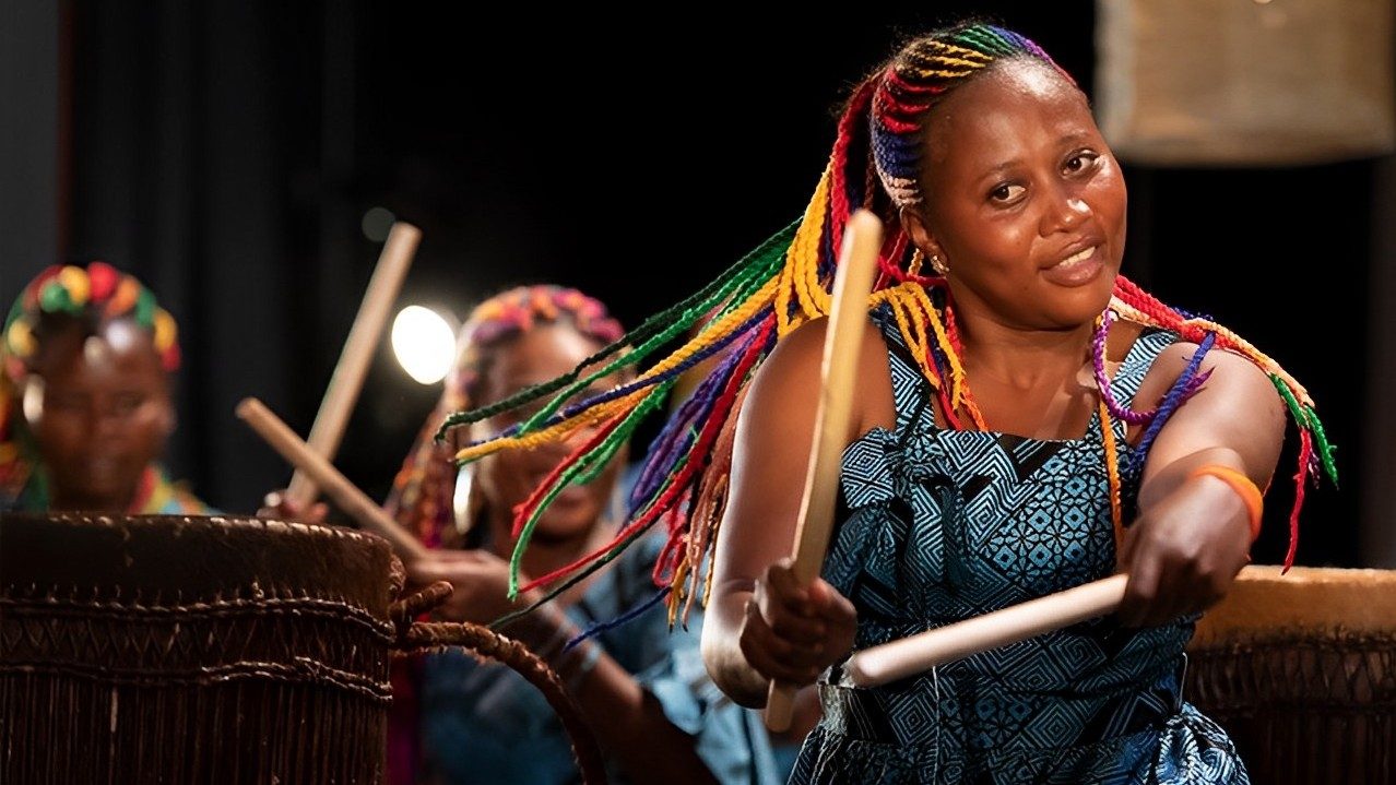  A Rwandan drummer performs "The Book of Life" on stage. She is a young Black woman with long, brightly colored braids, and she wears a blue and brown patterned dress.