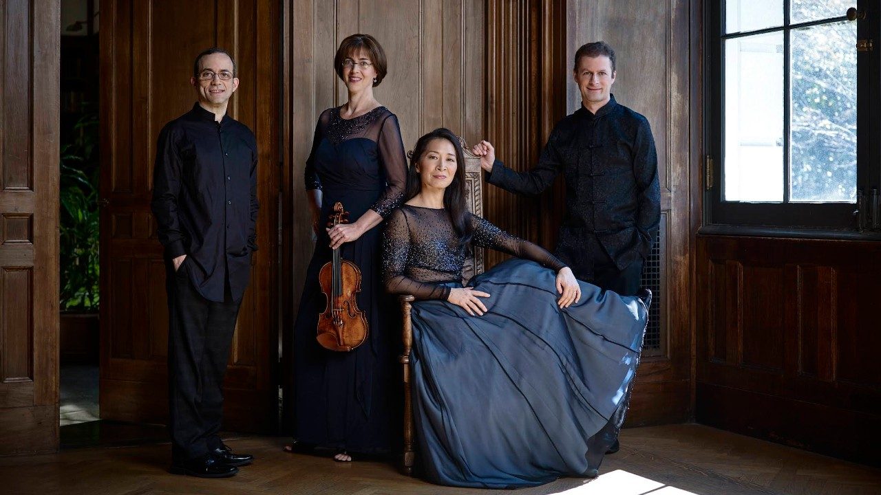  The Brentano String Quartet wears evening attire in a grand, wood-paneled room, light streaming in from large windows to the right. The quartet is comprised of two white men, one white woman, and one Asian woman, who is seated. The white woman holds her instrument, a violin.