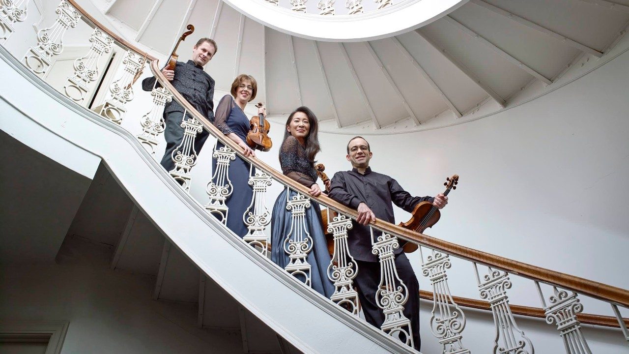  The Brentano String Quartet wears evening attire and poses on an ornate grand staircase that spirals up a foyer. The quartet is comprised of two white men, one white woman, and one Asian woman. They all hold their instruments and look down towards the camera.