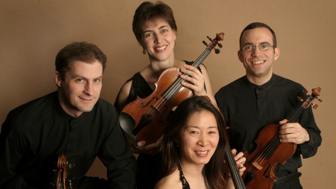  The Brentano String Quartet, from left, a white man, a white woman, an Asian woman, and a white man, wear black clothes and sit or stand close together, holding their instruments in front of a chai-colored background.
